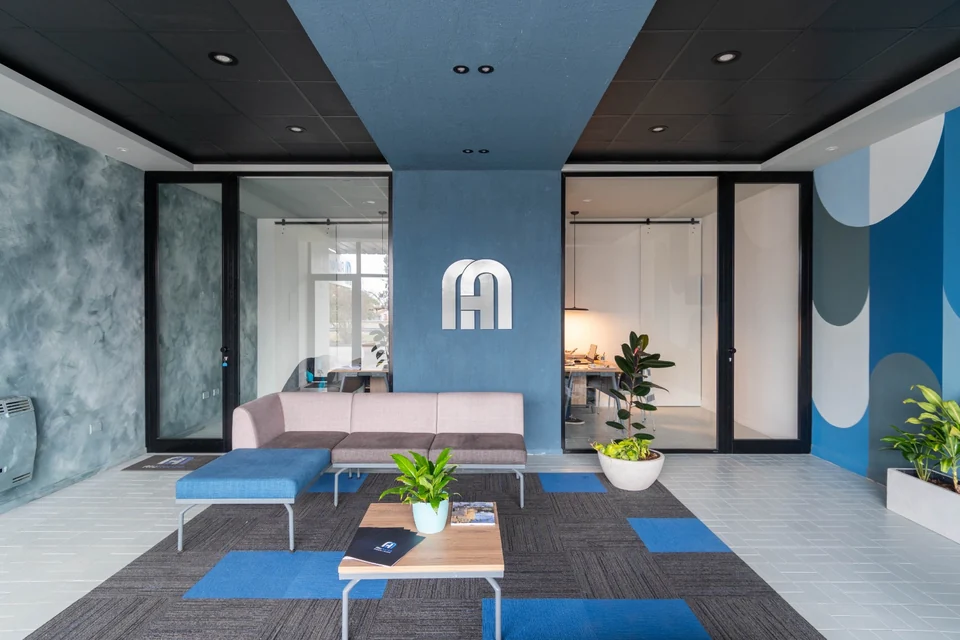 Reception with a blue plane crossing the black ceiling and folding down the wall, a metal arched monogram, glass and aluminium partitions, and an arch mural on the far wall