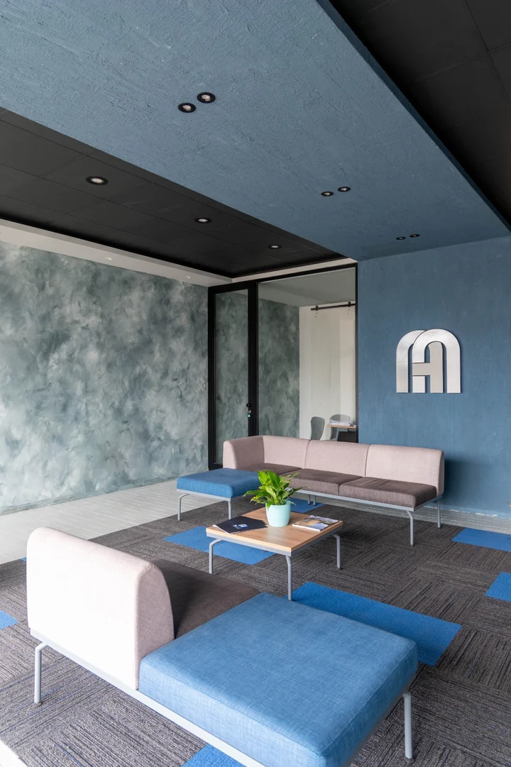 Seating area seen from the side, with the blue ceiling plane, the troweled green-grey wall and the metal monogram