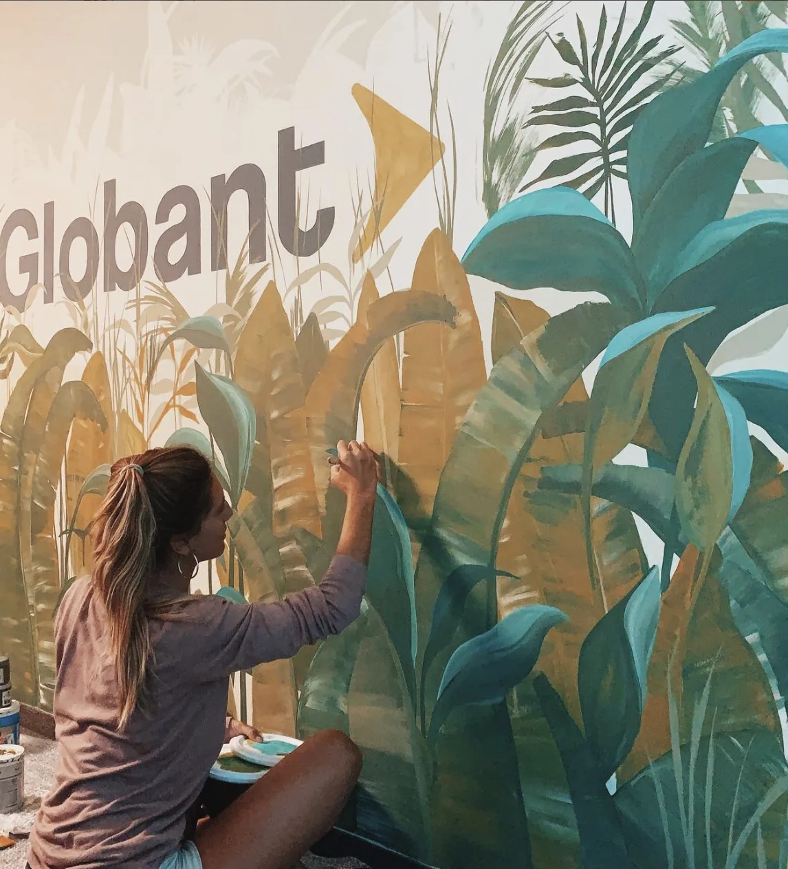 Belén Marcuzzi painting a large botanical mural on an office wall, with the Globant wordmark integrated into the foliage
