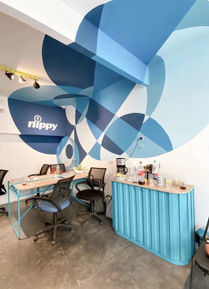 Office interior with a large geometric mural of overlapping circles in blues, wrapping across the wall, the corner and the underside of a beam, with the Nippy wordmark painted in the darkest area