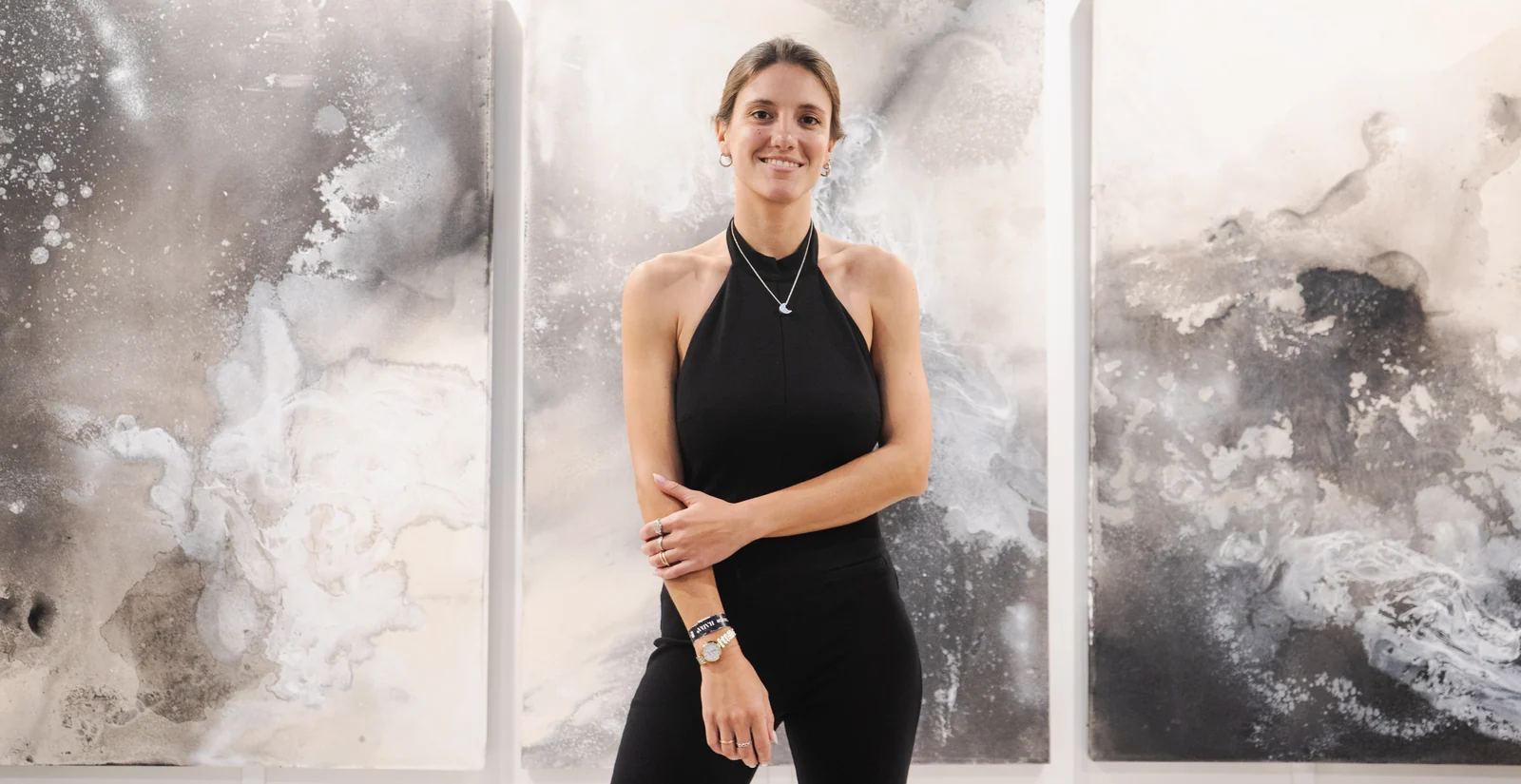 Belén Marcuzzi at her art-fair booth, standing in front of three large canvases in greys, whites and earth tones, under a sign with her name